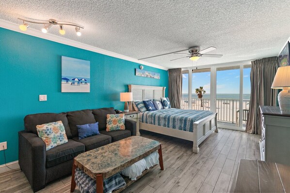 A room with a bed, a sofa, a small table, and a balcony with a view of the beach.