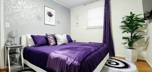 Un dormitorio con cama, mesita de noche, lámpara, planta en maceta y ventana con cortinas.