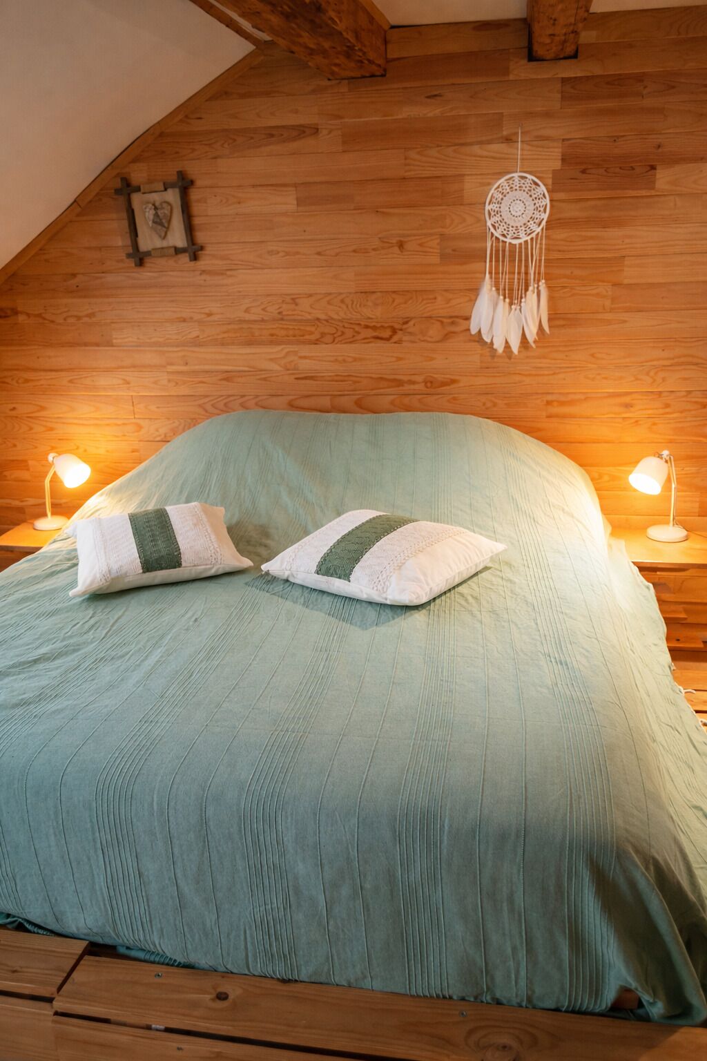 A bedroom with a wooden ceiling, a bed with a green bedspread, two bedside lamps, and a dreamcatcher on the wall.