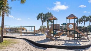A playground with slides and swings, surrounded by palm trees and a clear sky. - Branch Breeze - Golf Cart | Linens | Ocean Lakes (Myrtle Beach)