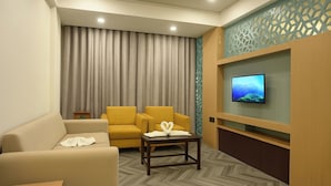 A hotel room with a sofa, two armchairs, a small table with a towel, a TV mounted on the wall, and a patterned herringbone floor.