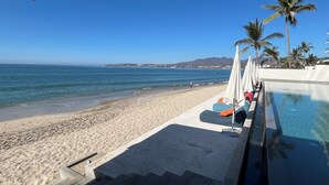 A beachside pool area with lounge chairs and umbrellas, a sandy beach, and a clear view of the ocean and distant mountains.