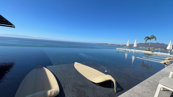 An infinity pool with clear water, lounge chairs, and a view of the ocean and mountains.