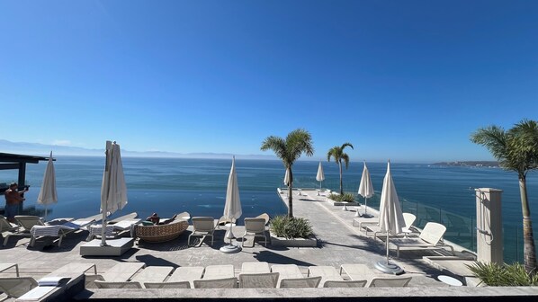 Un espace de détente extérieur, baigné de soleil, avec des chaises longues et des parasols, offrant une vue sur une mer calme et des montagnes lointaines.