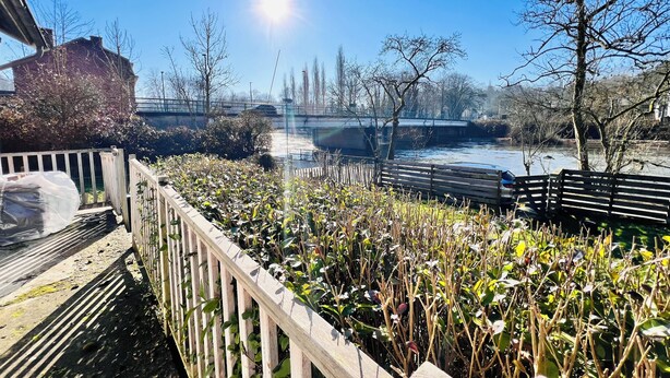 A riverside walkway with a wooden fence, a bench, and a bridge in the background. - Maison proche rivière avec jardin  (Esneux)