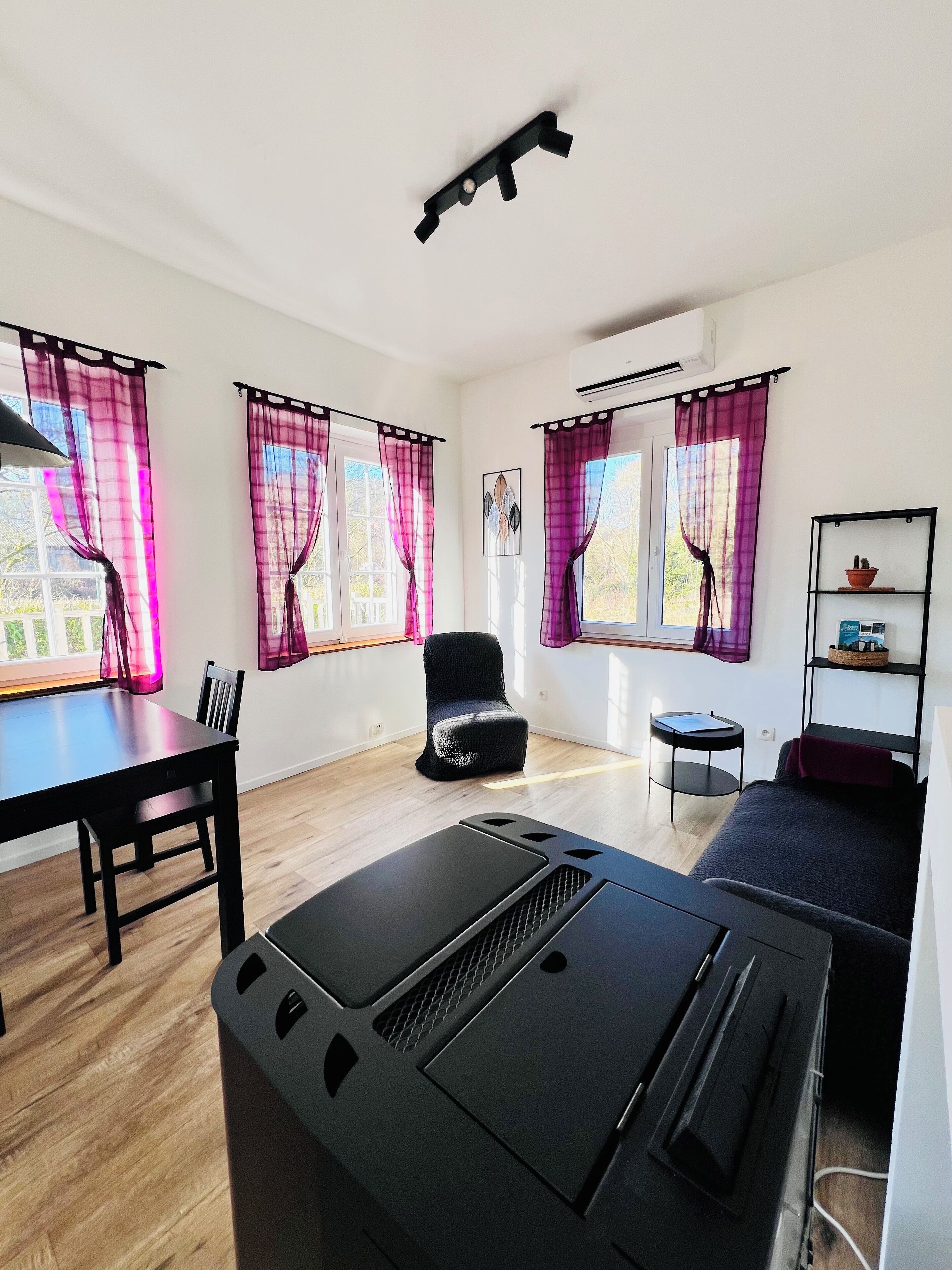 A modern, well-lit room with a dining table, a sofa, and a chair. There are large windows with purple curtains, a ceiling fan, and a small shelf with decorative items.