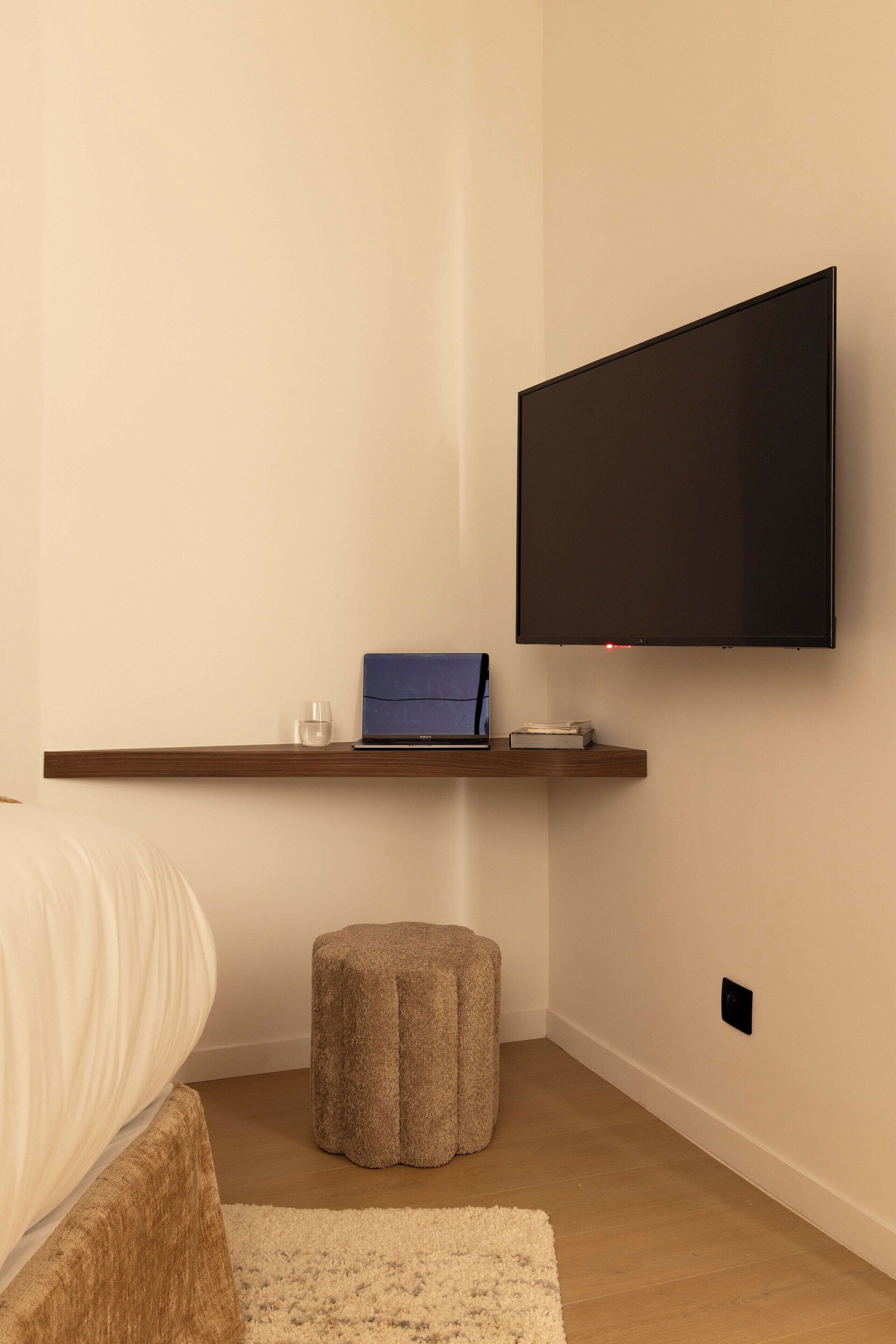 Un dormitorio moderno con una cama, una mesita de noche, un taburete pequeño, un televisor de pantalla plana montado en la pared y un estante con una computadora portátil y un vaso.