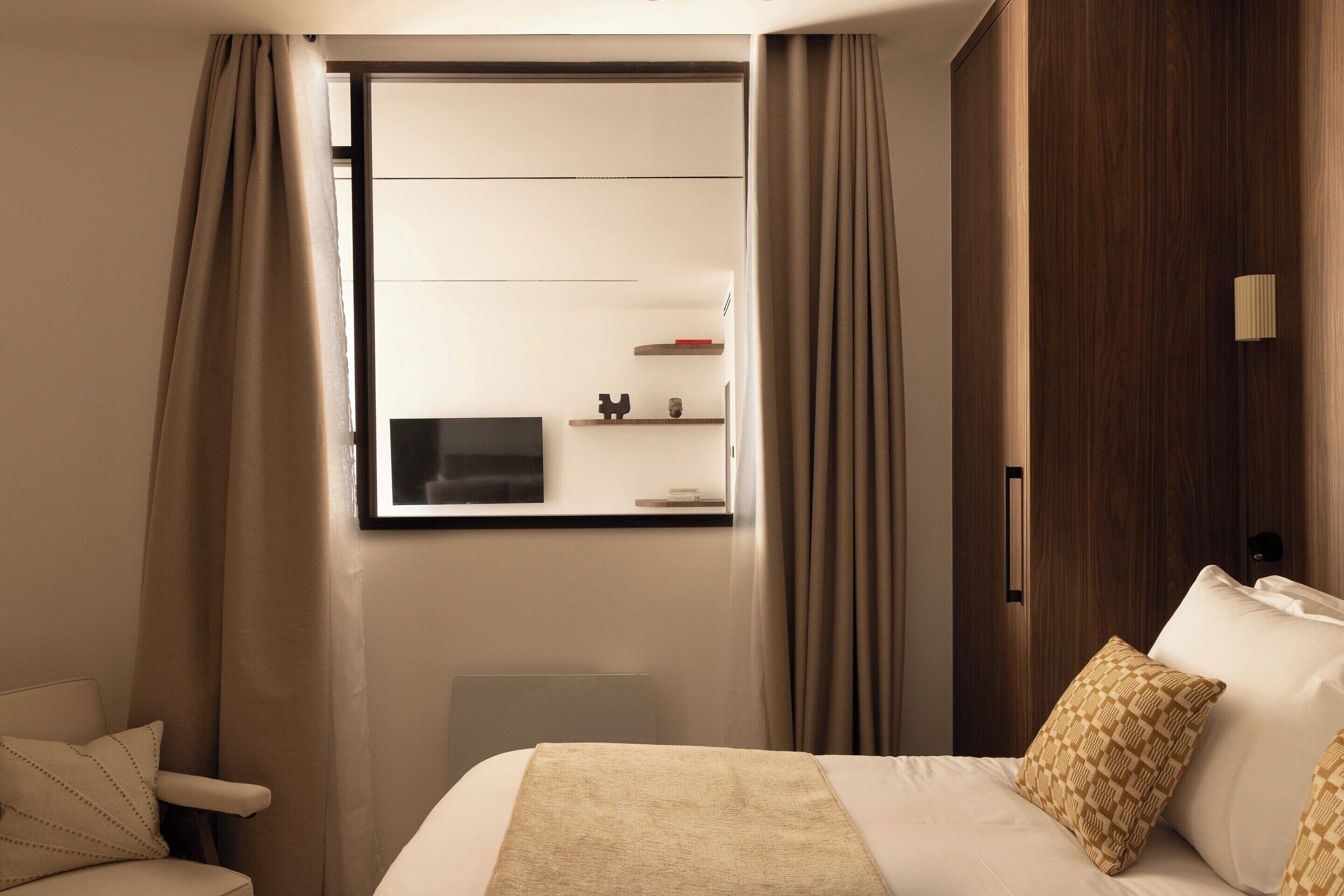 A hotel room with a bed, a chair, a window with curtains, and a wooden wardrobe.