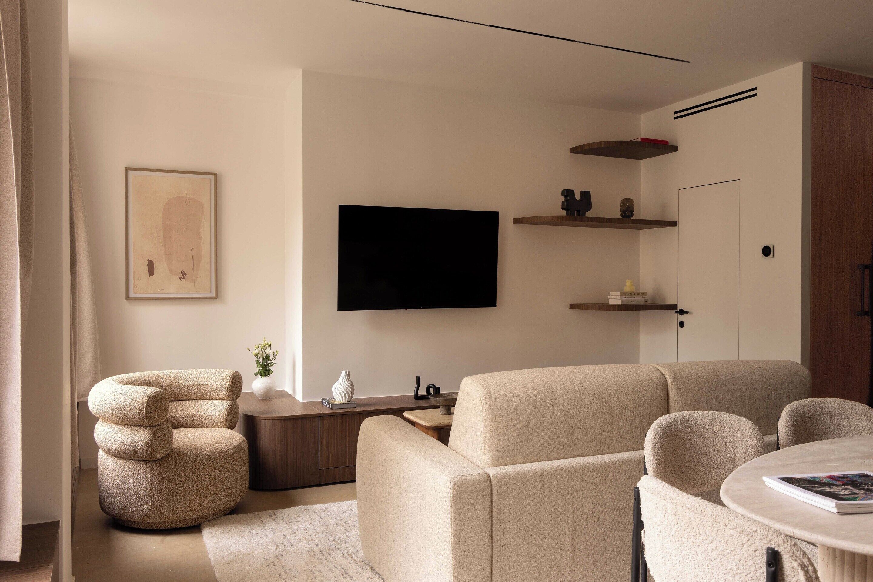 A modern living room with a sofa, armchair, and a round coffee table. There is a flat-screen TV mounted on the wall and a small side table with decorative items.