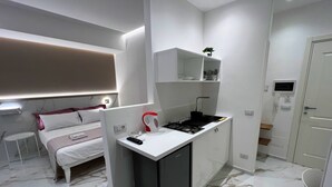 Quarto de hotel pequeno com uma cama, uma kitchenette compacta, uma televisão com moldura branca e um banheiro com chuveiro e vaso sanitário.