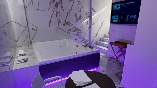 Casa de banho moderna com parede de mármore, banheira, uma pequena mesa com toalhas e uma televisão a exibir um filme.