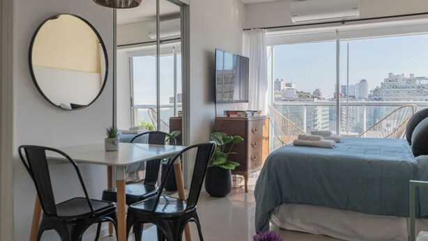 A modern bedroom with a large bed, a dining table with chairs, a TV, and a balcony with a city view. - MOT Libertador 5700 (Buenos Aires)