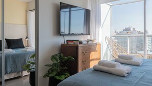A modern hotel room with a large bed, a TV mounted on the wall, a wooden dresser, and a view of the city through large windows. - MOT Libertador 5700 (Buenos Aires)