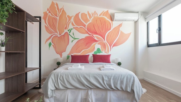 A hotel room with a large bed, a wooden floor, a wall mural of flowers, a shelf with plants, and a window with a view. - MOT Libertador 6300 (Buenos Aires)