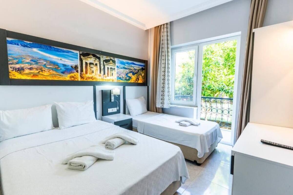 A hotel room with two beds, a desk, a nightstand, and a large window with a view of greenery.