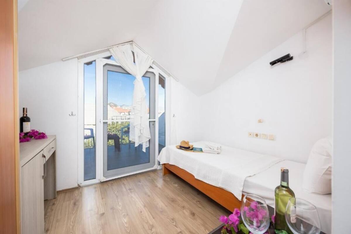 A modern bedroom with a large sliding glass door leading to a balcony, a white bed, a small desk, and a bottle of wine on the nightstand.
