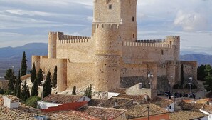 A historic stone castle with a central tower, surrounded by terracotta-tiled rooftops and cypress trees. - Cervantes Apartment (Villena)