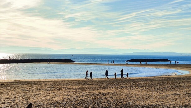 A beach with people walking and standing, a clear sky, and distant mountains.