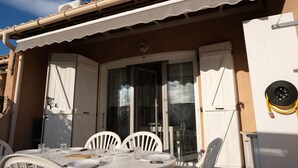 An outdoor dining area with a table set for two, white chairs, and a glass door leading to a covered patio.