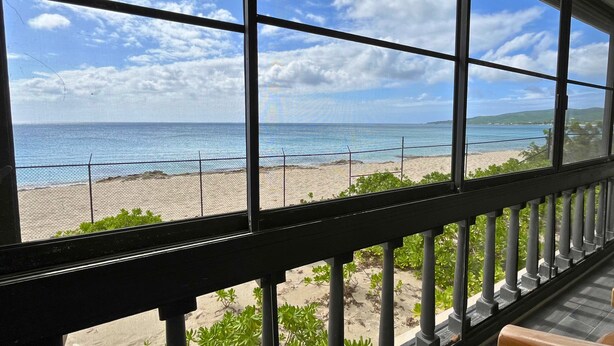 A view from a balcony with a metal railing, overlooking a beach with a fence and greenery. - Leisure Daze by the Sea (Frederiksted)
