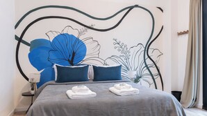A neatly made bed with a grey bedspread and two blue pillows, a wall mural with floral and abstract designs, and a small bedside table with a lamp and a potted plant. -  MOT Libertador 6300 A (Buenos Aires)