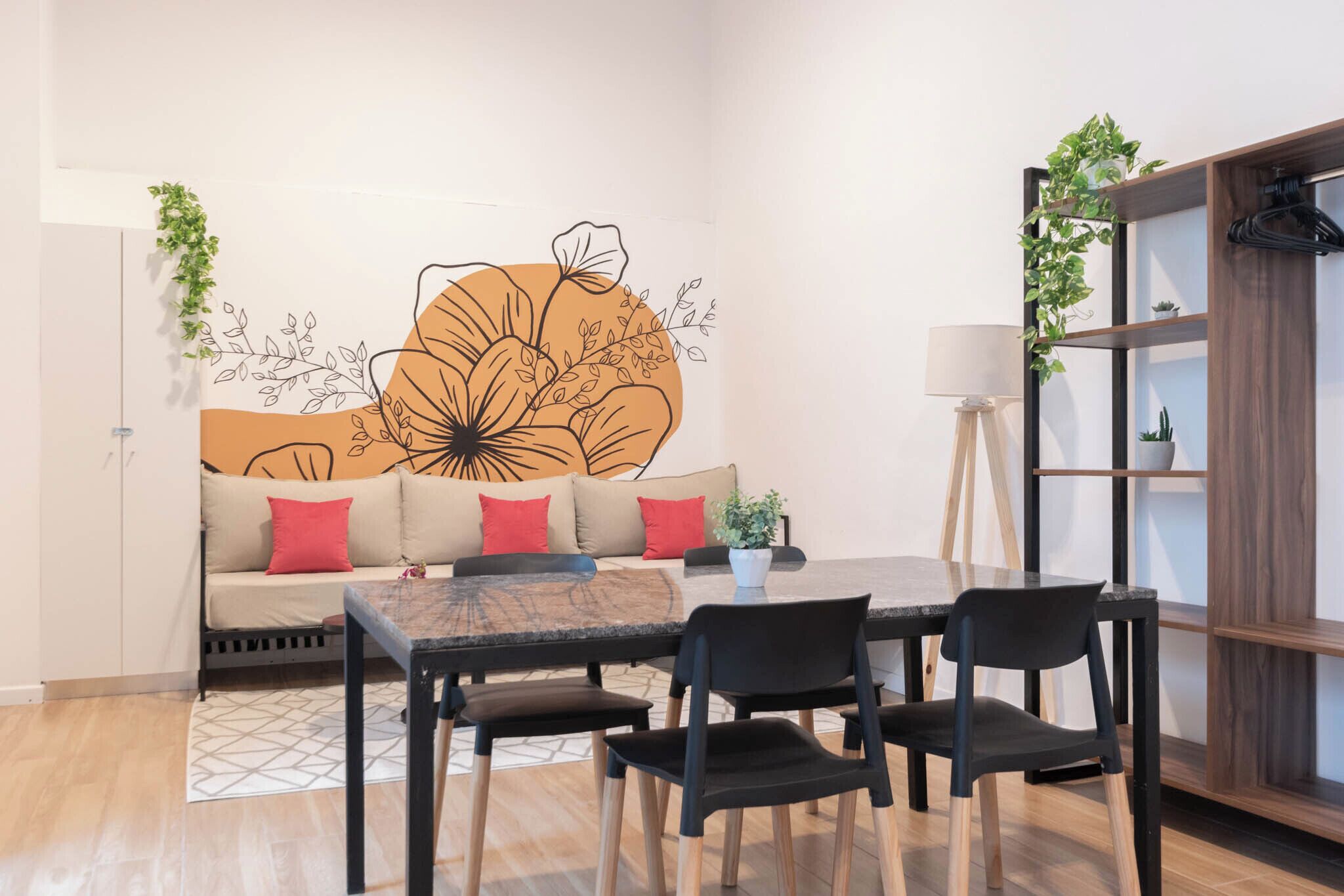A modern living room with a sofa, a dining table, and chairs. There is a wall mural, a floor lamp, and a shelving unit with plants.