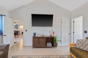 A living room with a flat-screen TV, a wooden cabinet, a sofa, and a dining area in the background.