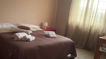 Cama bem arrumada com travesseiros e toalhas enroladas, mesa de cabeceira com abajur, quadro na parede e janela com cortinas.