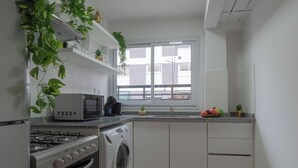 Une cuisine moderne équipée d’une cuisinière à gaz, d’un four à micro-ondes et d’un lave-linge, avec des meubles blancs et une fenêtre donnant sur des immeubles.