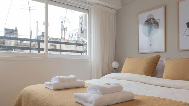 Une chambre d’hôtel avec un lit, deux serviettes blanches, une fenêtre donnant sur des immeubles et deux tableaux encadrés accrochés au mur.