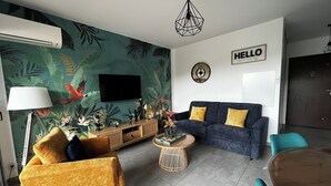 Ein Wohnzimmer mit tropischem Wandpapier, einem blauen Sofa mit gelben Kissen, einem Holztisch und einem flachbildfernseher, der an der Wand befestigt ist.