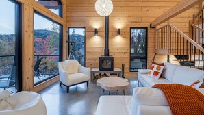 A cozy cabin interior with a fireplace, wooden walls, and large windows offering a scenic view. - Harfång - Spectacular view of Mount Edward (L'Anse-Saint-Jean)