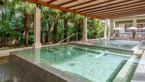 A pool area with two pools, palm trees, and lounge chairs under a wooden pergola.