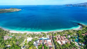 An aerial view of a coastal resort with a large pool, beachfront, and lush greenery.