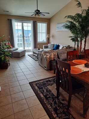 A living room with a sofa, a dining table with chairs, a ceiling fan, and a large window with curtains.