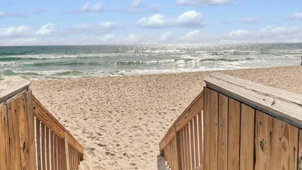 Una pasarela de madera que conduce a una playa arenosa con olas del océano al fondo.