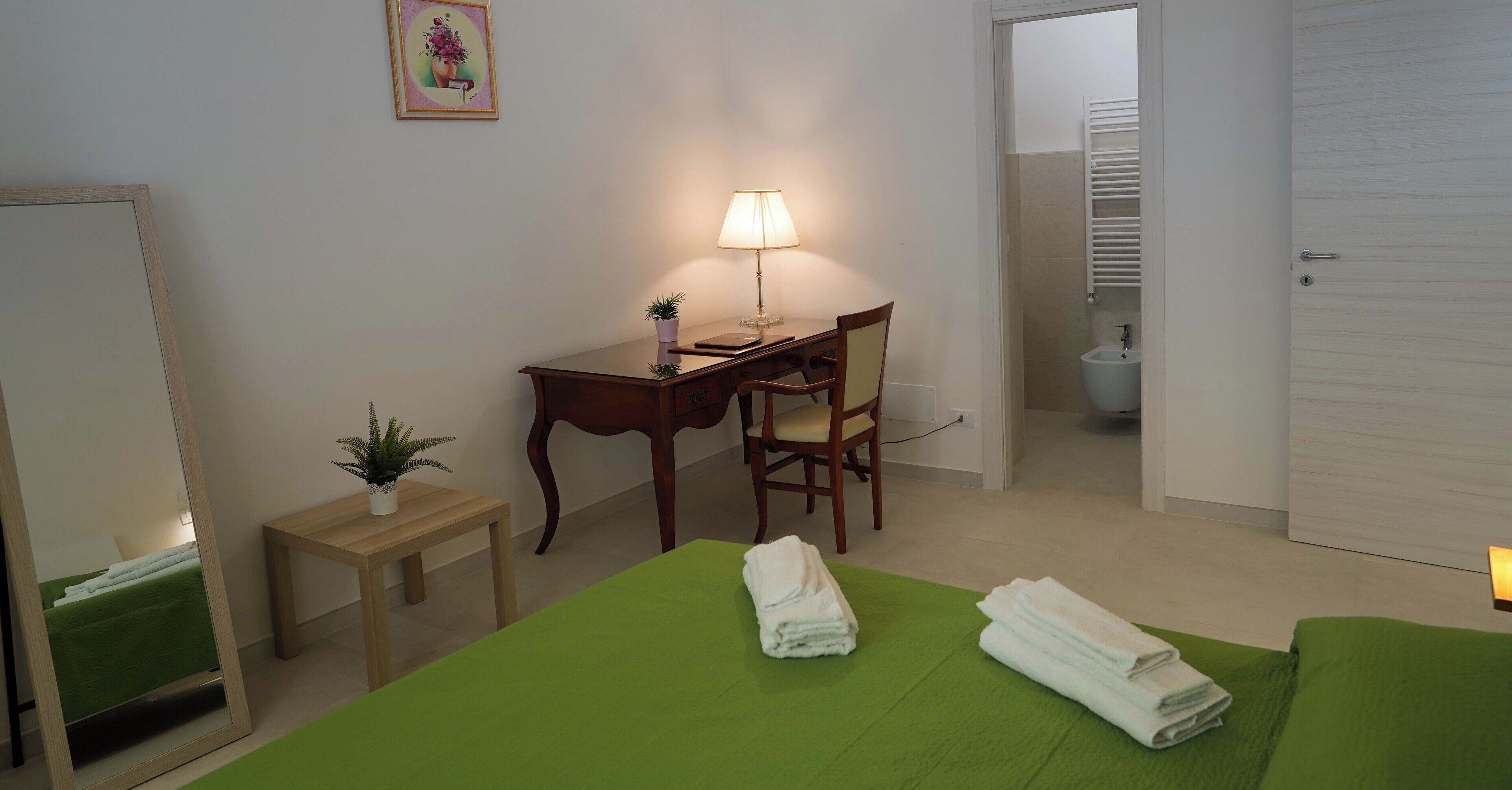 A hotel room with a bed, a desk, a chair, a small table with a plant, and a bathroom visible through an open door.