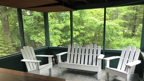 A covered outdoor seating area with a wooden table and white Adirondack chairs, overlooking a forested area. - Cozy Lakeside Escape on Lake Pleasant. (Lake Pleasant)