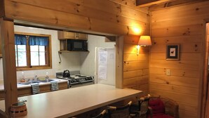 A wooden cabin interior with a kitchen featuring a stove, microwave, and refrigerator. There are two bar stools and a red chair. - Cozy Lakeside Escape on Lake Pleasant. (Lake Pleasant)