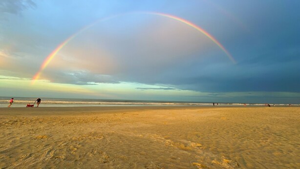 Een strand met een regenboog, zand en mensen in de verte.