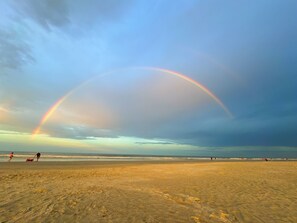 A beach with a rainbow, sandy terrain, and people in the distance.