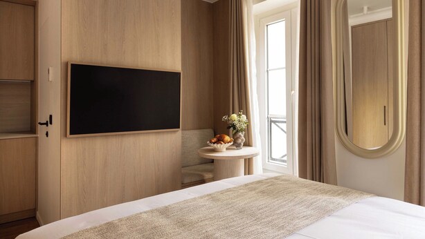 A hotel room with a bed, a flat-screen TV mounted on the wall, a small round table with a vase of flowers, and a mirror with an oval frame. - Charming Studio With AC - 2P- Musee du Louvre (Paris)