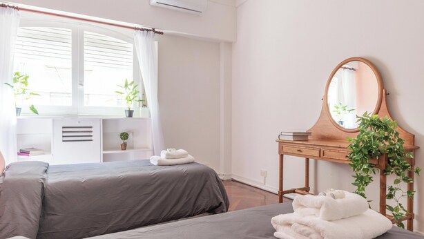 Quarto com duas camas, cómoda de madeira com espelho, planta em vaso e janela com cortinas.