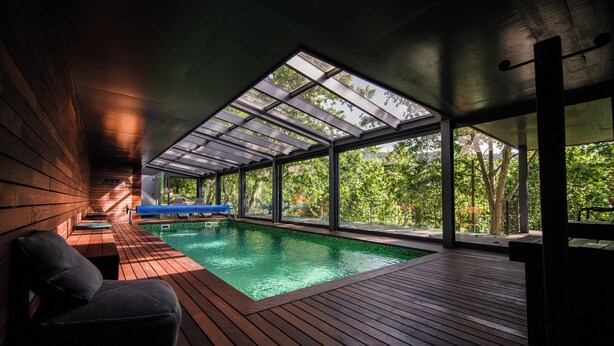 Piscina interior moderna con solárium de madera y grandes ventanales.