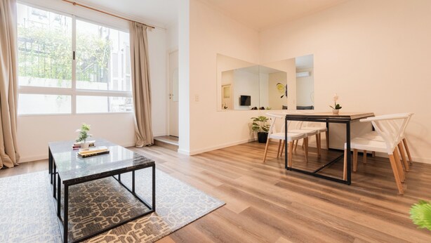 Una sala moderna con una mesa de centro de vidrio, una mesa de comedor con sillas, un ventanal con cortinas y un espejo en la pared.