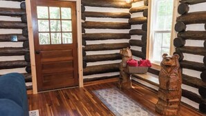 Interior de una cabaña de troncos con una puerta de madera, un banco con una cesta y dos esculturas de osos.