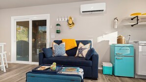 A living room with a dark blue sofa, a small ottoman, and a teal-colored coffee table. There is a white air conditioning unit on the wall, a small teal refrigerator, and a white cabinet.