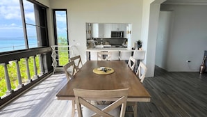 A dining area with a wooden table and chairs, a view of the ocean, and a kitchen in the background. - Green Flash by the Sea (Frederiksted Southeast)