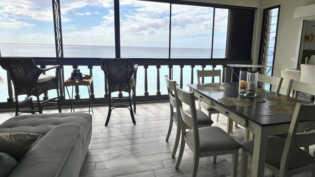 A dining area with a view of the ocean, a ceiling fan, and a balcony with chairs. - Green Flash by the Sea (Frederiksted Southeast)