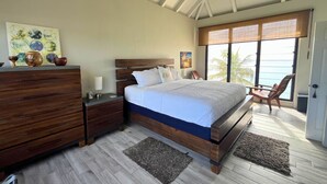 A bedroom with a wooden bed, a nightstand, a dresser, and a large window with a view of the sea. - Green Flash by the Sea (Frederiksted Southeast)